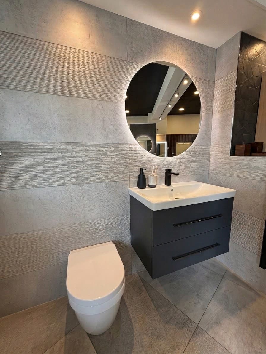 A cloakroom set in the showroom: ribbed and stone-effect grey tiling, a round backlit mirror over a charcoal wall-hung vanity, and a wall-hung WC